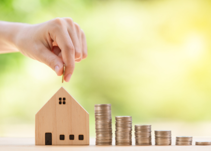 A hand is placing a coin into a wooden house-shaped piggy bank. Beside the house, there are stacks of coins arranged in ascending order, suggesting growth or savings. The background is a blurred green and yellow, indicating an outdoor, natural setting.