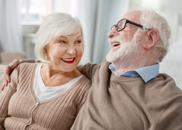 An elderly couple is sitting close together, smiling and laughing. The woman on the left is wearing a light brown cardigan over a white top and has short white hair. The man on the right is wearing black-framed glasses, a light blue collared shirt, and a beige sweater. Both appear to be happy and enjoying each other's company. The background features soft white and light tones, with sheer curtains and a window letting in some natural light.