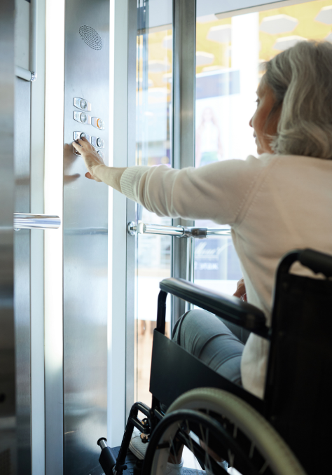 An elderly person with gray hair is sitting in a wheelchair inside an elevator, reaching out to press a button on the control panel. The scene is well-lit, with reflections visible on the elevator's glass doors.