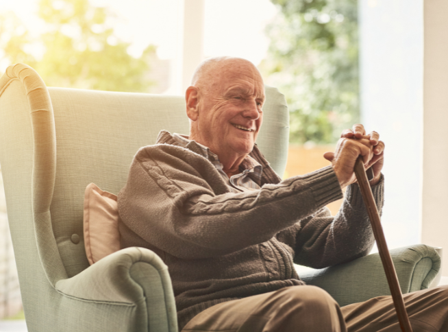 An elderly man is sitting comfortably in a light-coloured armchair, smiling and holding a walking cane with both hands. The sunlight is streaming in from a window behind him, illuminating the room and creating a warm, cozy atmosphere. He is wearing a grey sweater and light-coloured pants, and his bald head is reflecting the light subtly. There is a green, leafy background visible through the window.