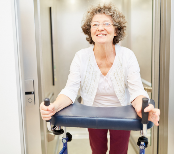 An elderly woman with curly gray hair and glasses is smiling while standing in an elevator with her walker. She is wearing a white cardigan over a light shirt and maroon pants. 