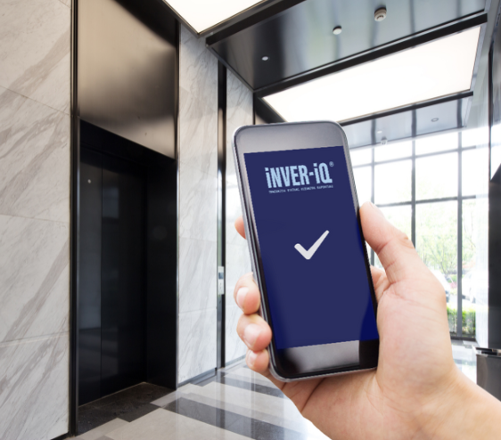 A person holding a smartphone in a modern lobby area. The phone screen displays the text "iNVER-iQ" with a checkmark below it. In the background, there are elevator doors and large windows letting in natural light. The environment is well-lit and the walls have a marble finish.