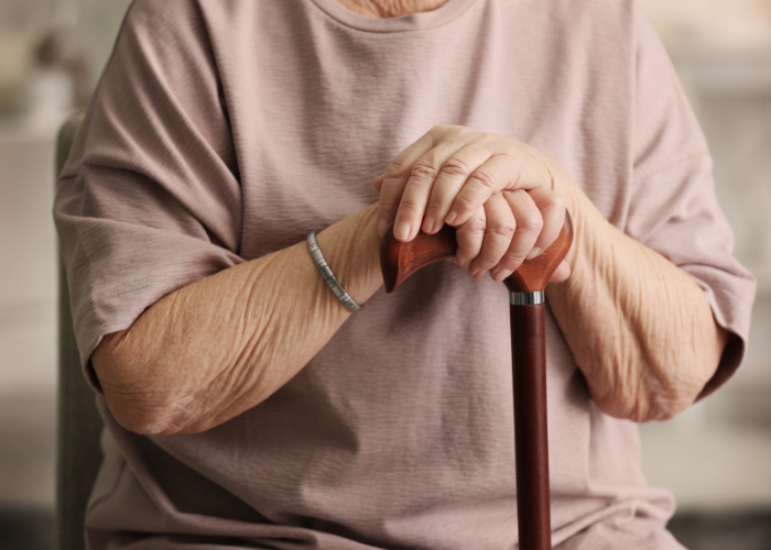 Close-up of an elderly person wearing a pink shirt, sitting and resting their hands on top of a wooden cane. The person is also wearing a silver bracelet on their left wrist.