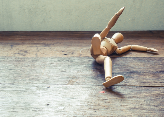 A small wooden artist mannequin is lying on a wooden floor with its limbs splayed in different directions, giving the appearance of having fallen or tripped. The background features a plain wall with a textured surface.