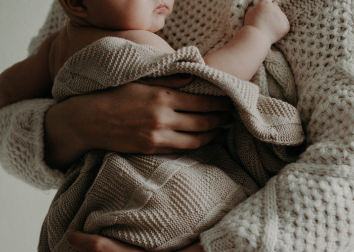 A close-up of a person wearing a cream-colored knit sweater, holding a baby wrapped in a soft, beige knitted blanket. The person's arms securely cradling the baby, providing a sense of warmth and comfort. The baby's face is partially visible and resting against the person's chest.