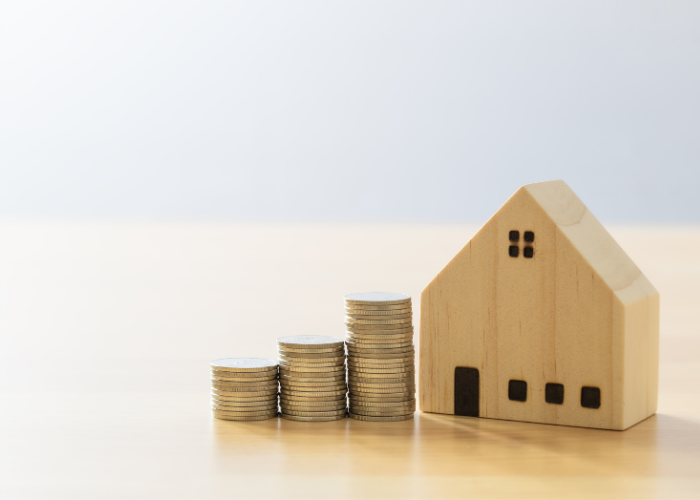 A small wooden house figurine is placed on a light wooden surface next to three stacks of coins arranged in increasing height. The background is plain and softly lit, emphasizing the concept of saving money or increasing wealth in relation to housing or real estate.