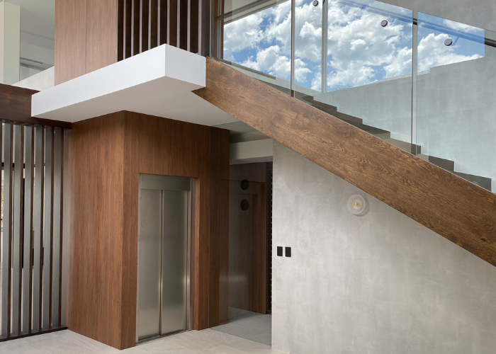 A modern interior space featuring a stainless steel elevator with a surrounding wooden frame. A sleek staircase with wooden steps and glass railing is positioned diagonally above the elevator. The walls are light gray, and the ceiling has a section with recessed lighting. The stairwell area offers a clear view of a bright, blue sky with scattered clouds through large windows. The design elements showcase a combination of wood, glass, and metal, creating a contemporary look.