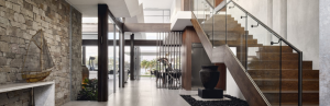 Elegant interior with a stone accent wall, modern staircase with glass railings, and large glass windows providing a view of lush greenery and the outdoors.