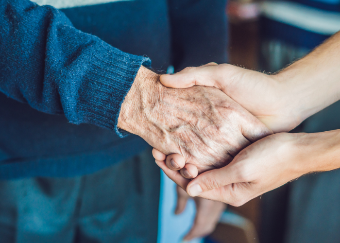 A close-up of a handshake between an elderly person's hand and a younger person's hand, symbolising connection and support. The elderly individual is wearing a dark blue sweater. The background is softly blurred.