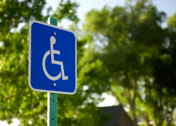 A blue sign with a white wheelchair symbol indicating accessible parking or facilities, set outdoors against a backdrop of green trees.