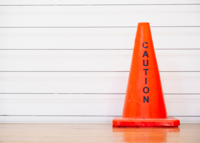 A bright orange caution cone with the word "CAUTION" written vertically in black letters, placed indoors on a wooden floor against a white wall with horizontal lines.