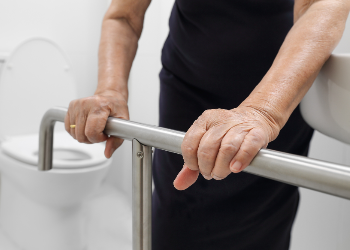 An elderly person grasping a stainless steel support rail in a bathroom, with a toilet visible in the background. The focus is on the hands and the rail, emphasising accessibility and safety.