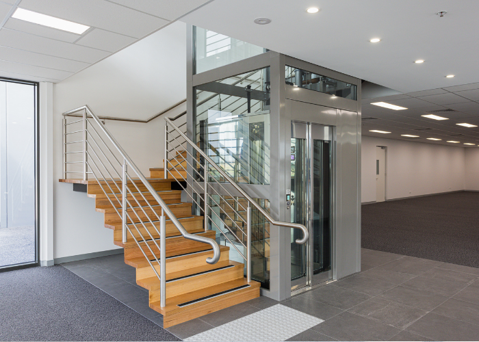 A contemporary interior featuring a staircase with wooden steps and metal railings next to a glass-walled elevator. The area has tiled flooring, with a spacious, carpeted open space visible in the background. Ample lighting comes from ceiling fixtures, and large windows allow natural light in.