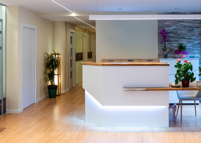 A modern reception area with a white desk and wooden countertop, featuring soft under-lighting. The space includes a potted plant and colorful flowers for decoration. The room has a wood floor, a stone accent wall, and a few doors leading to other areas.