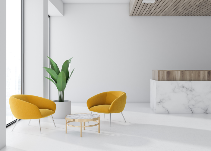 A minimalist lounge area with two mustard-yellow chairs and a round marble-top coffee table with gold legs. A large potted plant is placed beside the chairs. The room features large windows, a marble reception desk, and a wooden ceiling accent.