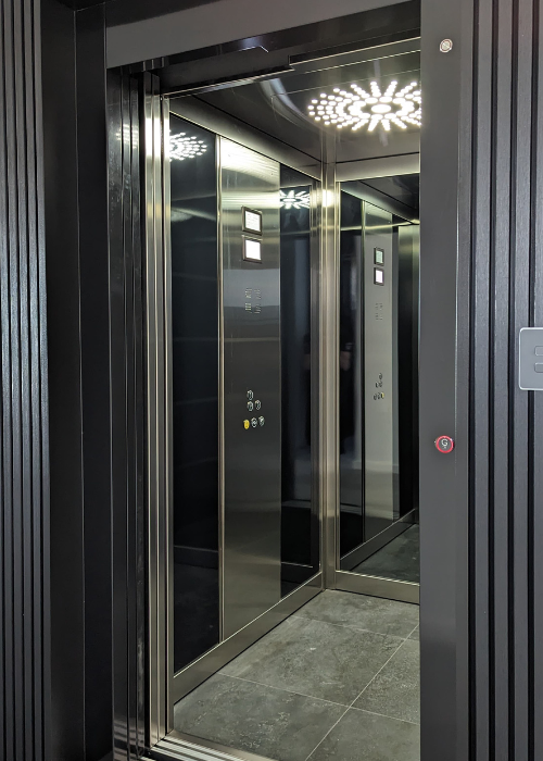 A modern elevator with open doors, revealing a sleek, reflective interior. The walls are lined with mirrors, and the ceiling features a decorative circular LED light. The floor is dark tiled. Control panels with buttons are visible on one side. The exterior frame of the elevator is matte black with subtle ridges on the walls.