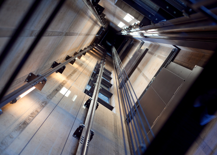 A view looking upward inside an elevator shaft. The vertical walls are lined with concrete, and several metal rails run alongside cables. Light shines from fixtures along the sides, illuminating the otherwise dim space. The image captures the depth and engineering complexity of the shaft.