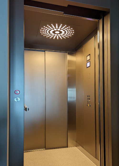 The interior of an open elevator with sleek metallic walls and ceiling. The ceiling features circular patterned lighting. The control panel on one side includes a digital display and numerous buttons. The floor is light, adding contrast to the metallic surfaces, and the overall design is modern and sophisticated.