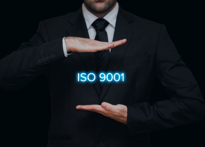 A man in a black suit holds his hands apart vertically, with the glowing text "ISO 9001" floating between them. The background is dark, highlighting the text and formal attire. The image conveys a sense of quality assurance and professionalism.