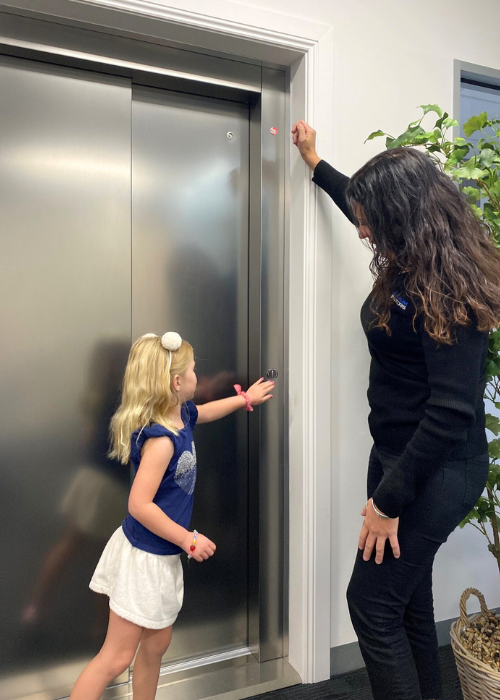 A woman and a young girl stand in front of a closed elevator with shiny metallic doors. The girl, wearing a blue shirt and white shorts, is reaching to press the elevator button. The woman, in black attire, is standing beside her, holding a pink object near the top of the elevator frame. A green plant decorates the area.