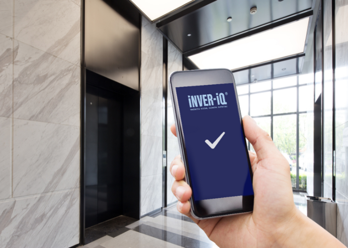 A person holds a smartphone displaying the "INVER-iQ" app with a large checkmark on the screen. The background shows a modern building lobby with marble walls and an elevator, suggesting successful access or operation using the app. Natural light enters through large windows.