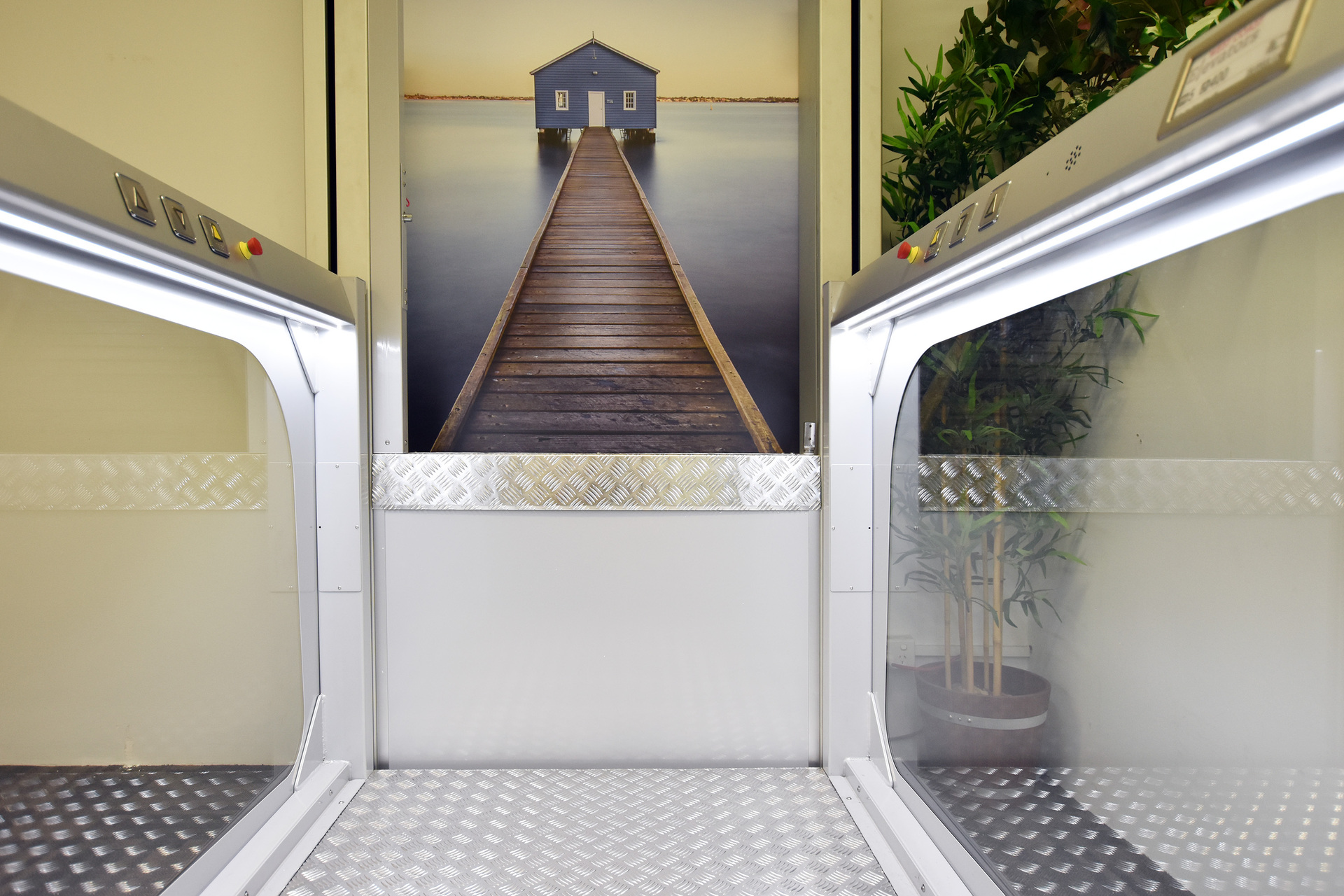 Interior of an elevator with stainless steel floors and control panel. A large wall mural depicts a long wooden pier leading to a small house over calm water.