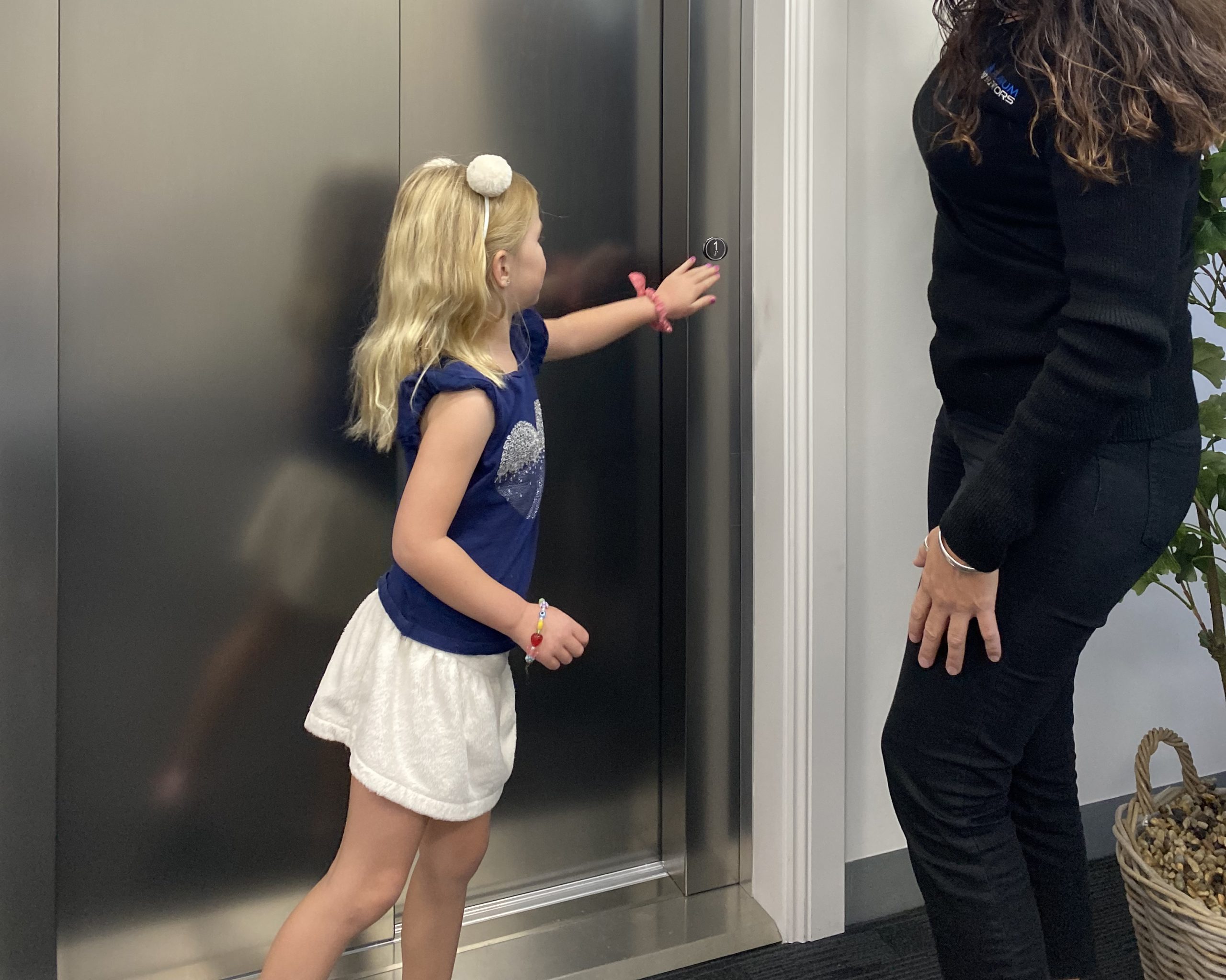 A young girl pressing an elevator button while standing next to an adult woman. They are in front of a stainless steel elevator door, with a plant visible to the side.