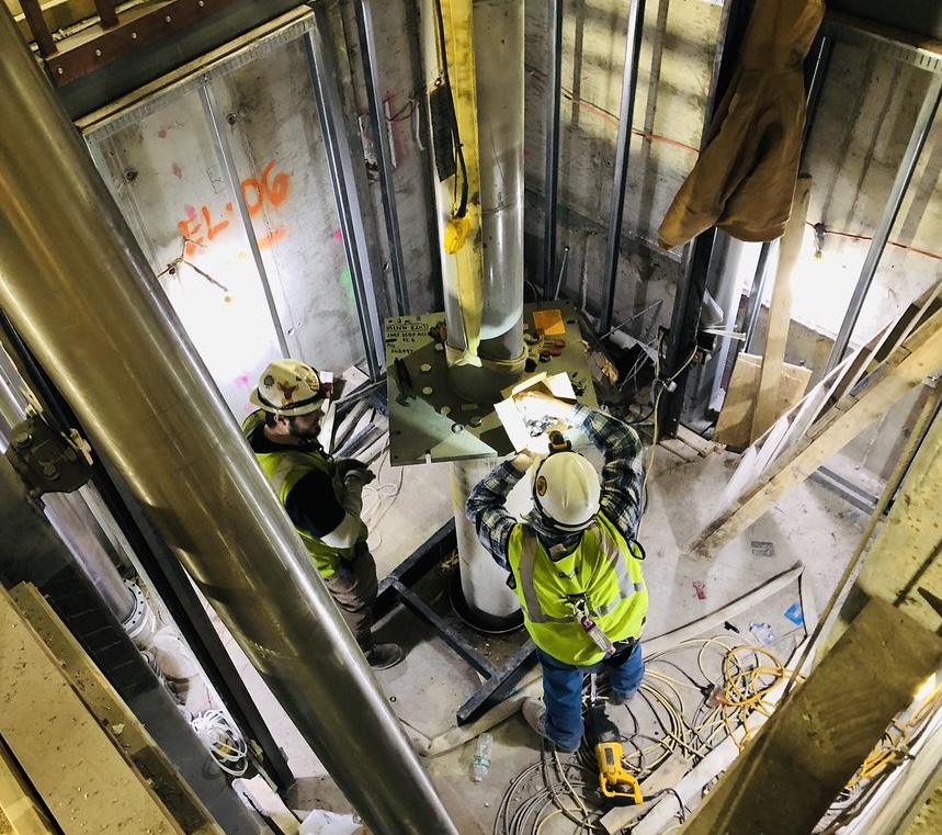 Construction workers inside an elevator shaft wearing safety gear and helmets. They are examining plans on a metal platform with tools and cables around them.