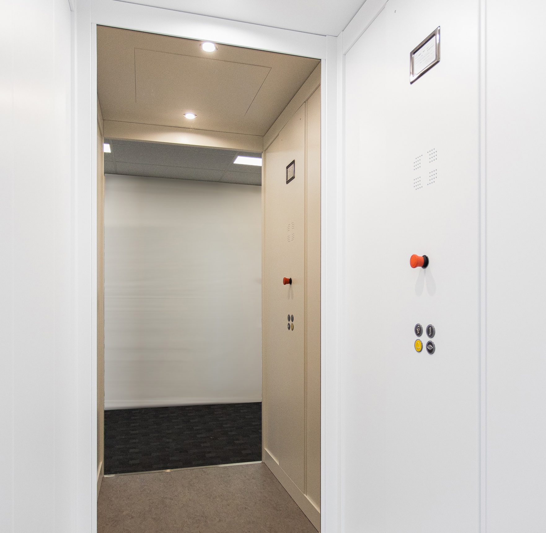 Interior of a clean, modern elevator with white walls and a simple control panel featuring buttons. The ceiling has integrated lighting, providing a bright atmosphere.