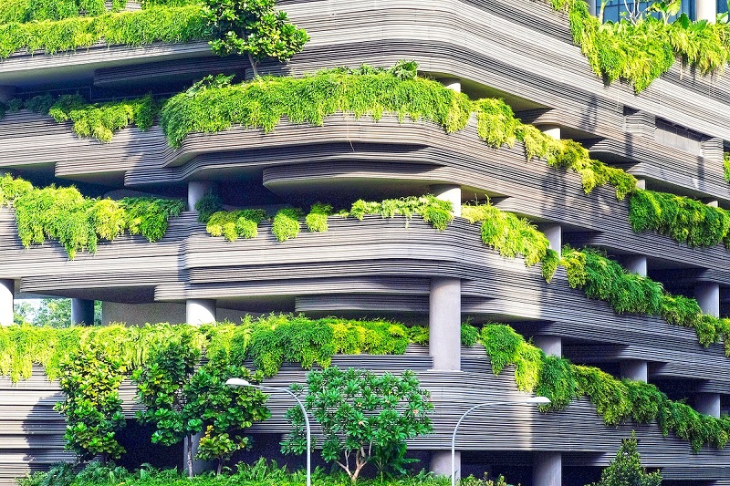 A vibrant, tiered building covered with lush greenery on each level. The structure features wavy, horizontal architectural layers with abundant plant life cascading over the edges. The light gray building contrasts with the bright green foliage, creating a dynamic blend of natural and urban design. Small trees and bushes are planted around the base, enhancing the structure's garden-like appearance.