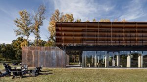 A modern building with a distinctive design featuring a rust-colored metal façade with horizontal slats. The structure includes large glass doors and windows, reflecting the surrounding greenery. In the foreground, there are outdoor black chairs arranged in small groups on a grassy area. Tall trees with autumnal foliage are visible in the background under a partly cloudy sky.