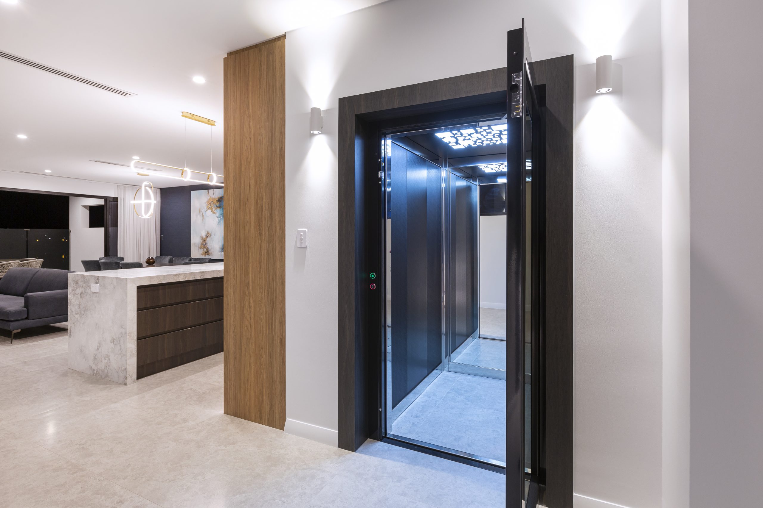 A modern, well-lit interior featuring a sleek elevator with glass and metal doors open to reveal its interior. The room has a luxurious design with beige tiled flooring, a marble kitchen island, and a seating area with dark gray sofas. Pendant lights hang from the ceiling, and artistic wall decor adds sophistication. The overall ambiance is contemporary and elegant.