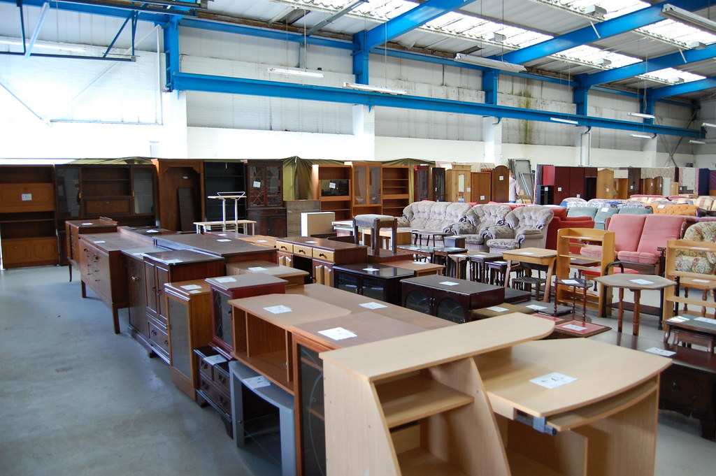 A large warehouse filled with assorted furniture, including wooden desks, cabinets, tables, chairs, and sofas of various styles and colours. The furniture is arranged in rows under a high ceiling supported by blue beams. The warehouse has bright overhead lighting and concrete flooring, creating an organised yet bustling atmosphere.