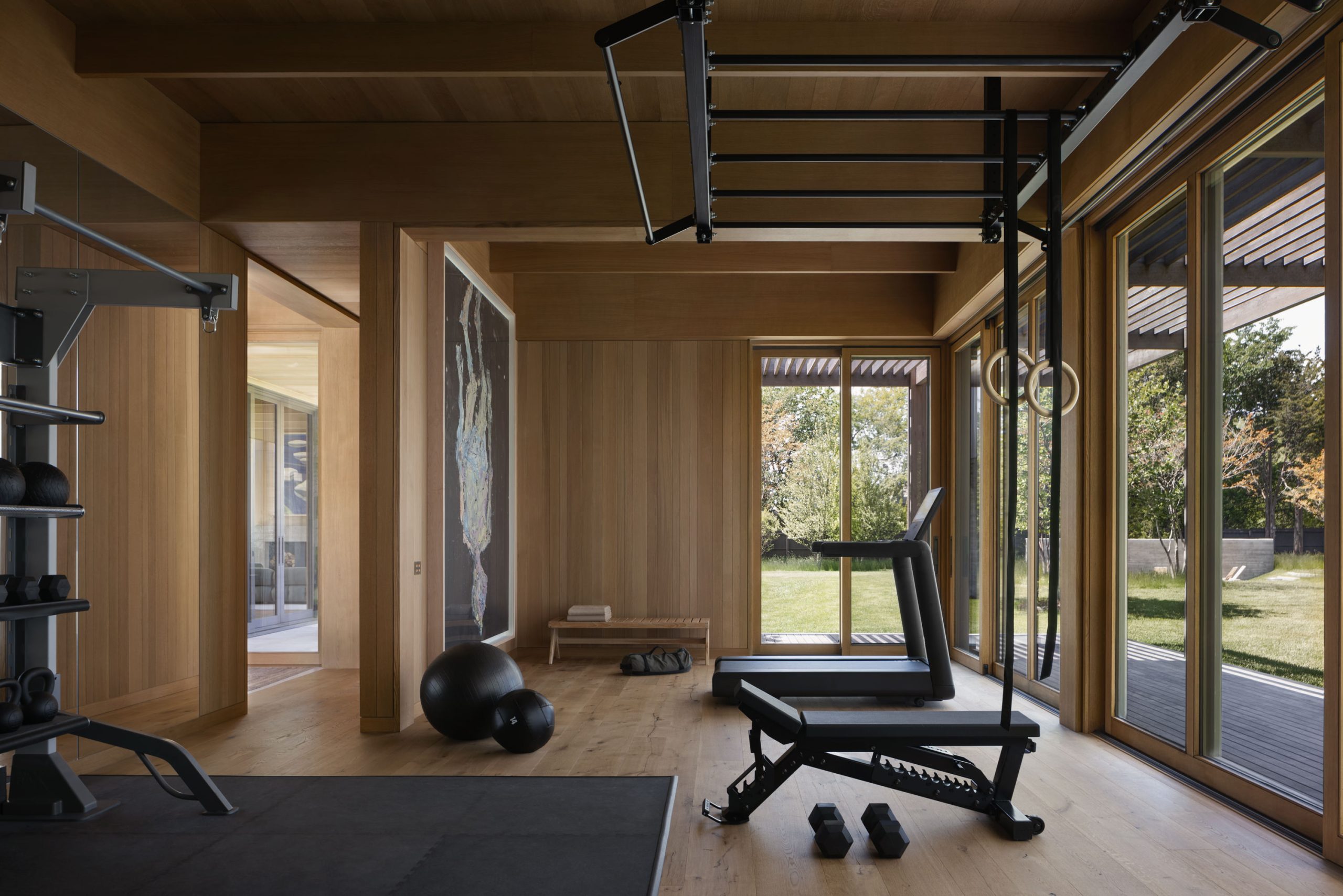 A modern home gym with wooden walls and floors, featuring large floor-to-ceiling windows that offer views of a green outdoor area. The gym is equipped with a treadmill, exercise rings, a bench, and free weights, with a rack for additional equipment. A yoga mat and exercise balls are positioned near a small wooden bench. The room has a spacious and airy feel, combining functionality with a natural aesthetic.