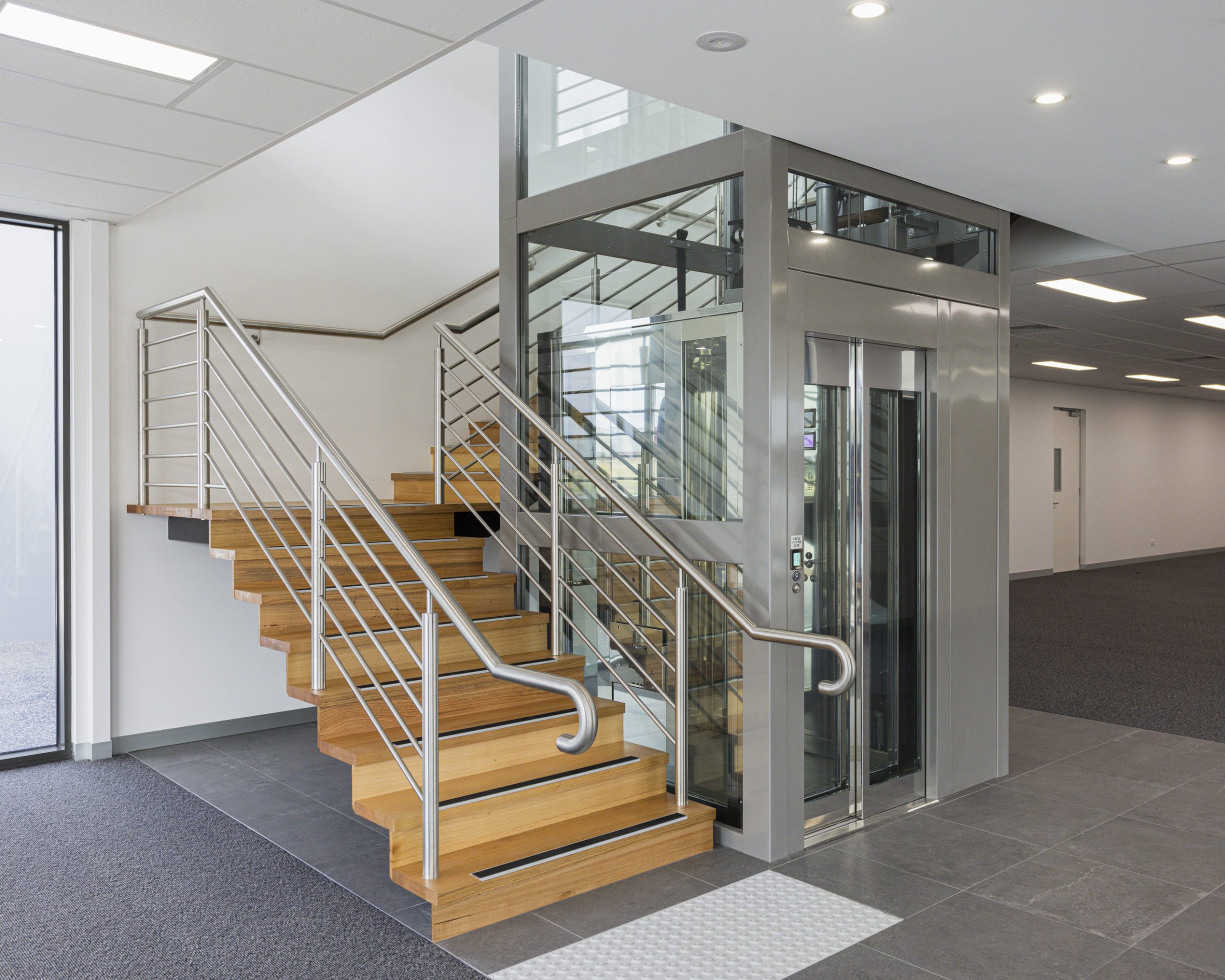 Modern staircase with adjacent glass elevator entrance.