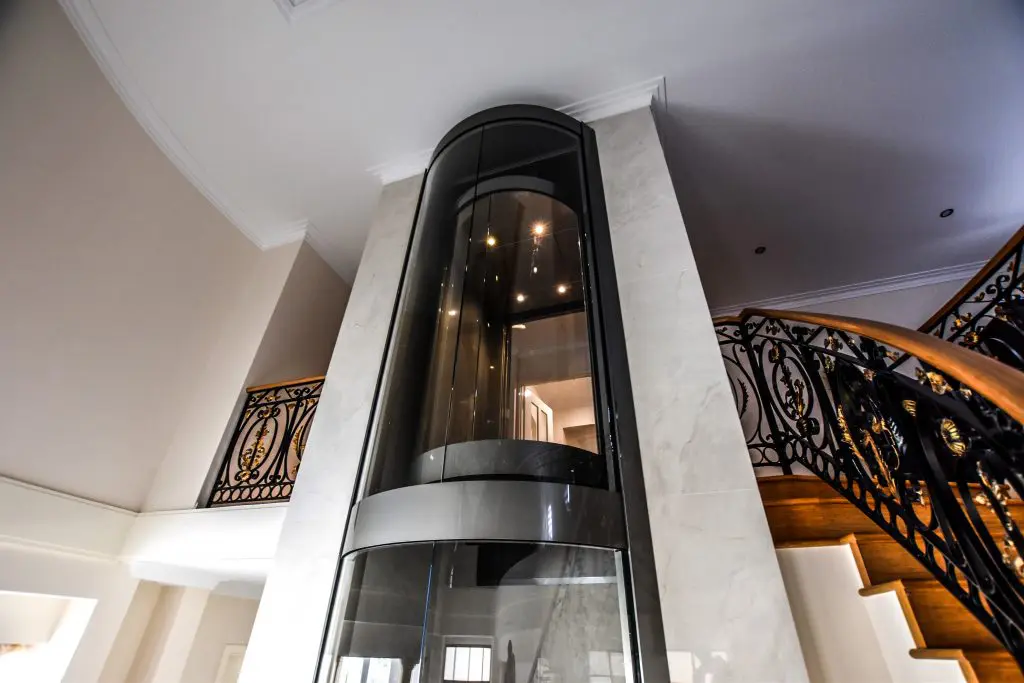 Luxury glass elevator beside ornate staircase in house.