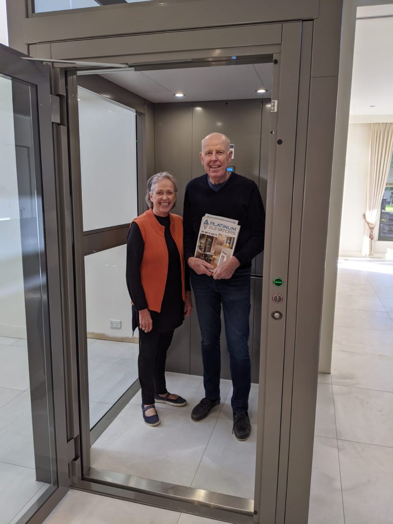 Handover celebration in Toorak - Sovereign Lift in Glazed Metal Structure Couple inside a home lift with brochure.